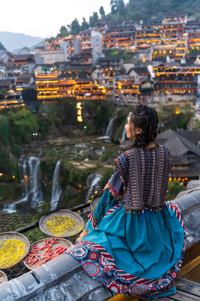 Mujer con traje tradicional frente al pueblo antiguo iluminado y las cascadas de Furong en China.