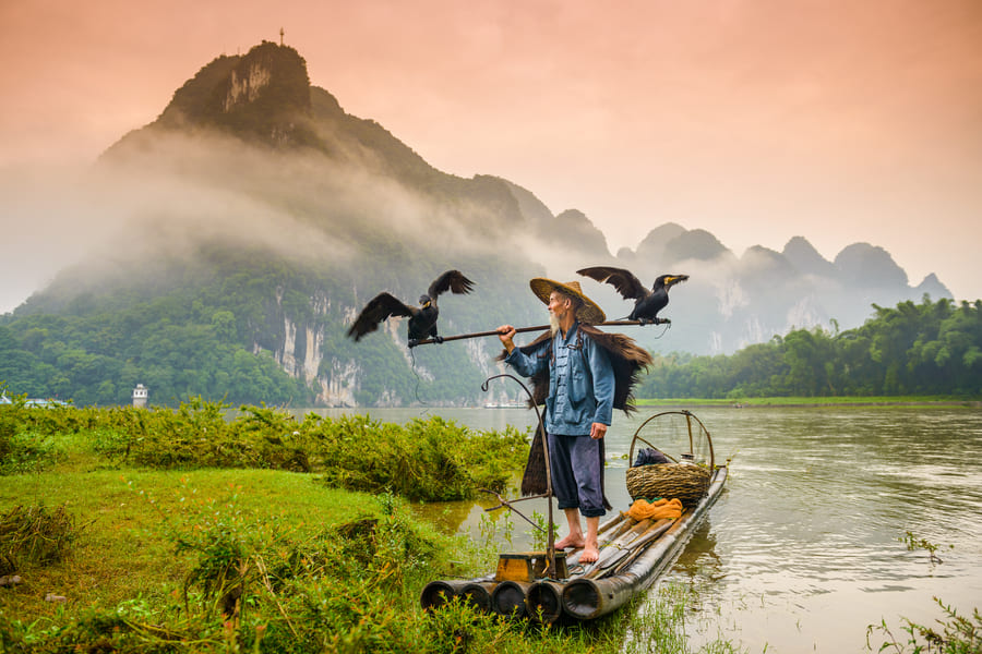Pescador de cormorán sobre una lancha en el río Yulong en Yangshuo con montañas de fondo
