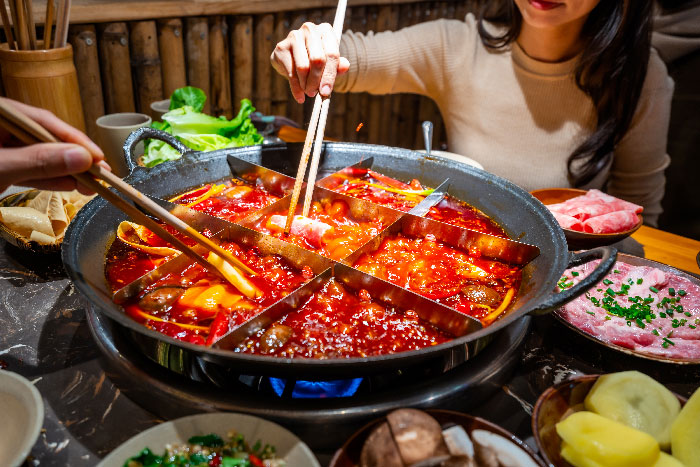 Personas utilizando palillos para comer un tradicional hot pot al estilo de Chongqing, mostrando un gran caldero dividido en compartimentos con un caldo rojo picante y burbujeante, rodeado de ingredientes frescos como carne y verduras.