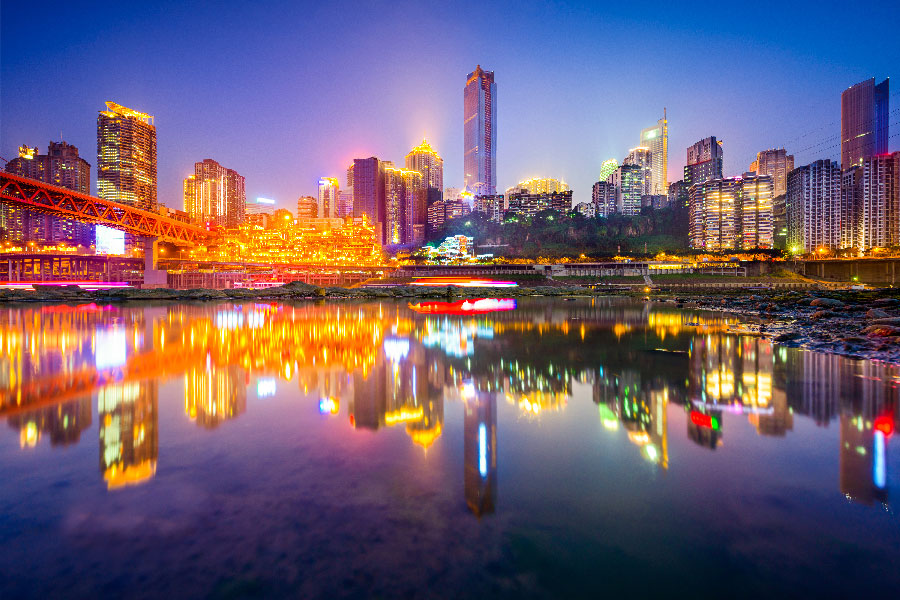 Skyline iluminado de la ciudad de Chongqing de noche, con rascacielos modernos reflejándose en las aguas tranquilas del río y un puente iluminado a la izquierda bajo un cielo crepuscular.