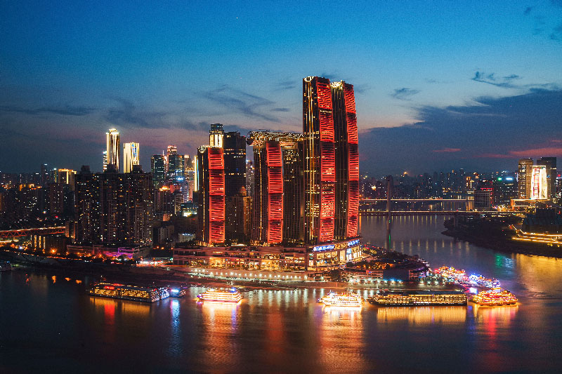 Vista nocturna del complejo Raffles City en Chongqing, destacando sus rascacielos iluminados con luces rojas en la intersección de los ríos, con barcos turísticos brillantes anclados en la orilla.