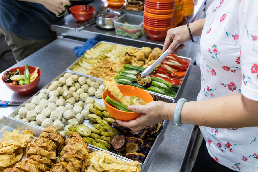 Variedad de ingredientes para Yong Tau Foo tradicional en un mercado callejero de comida asiática, con una persona sirviendo verduras rellenas en un tazón naranja.