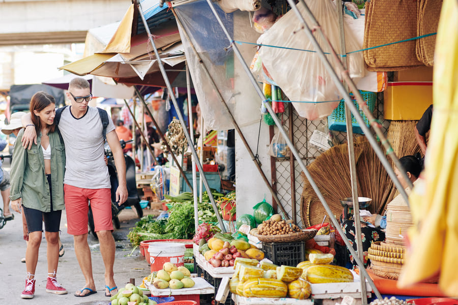Una joven pareja de turistas camina abrazada recorriendo los coloridos puestos de frutas y verduras en un mercado callejero asiático tradicional.