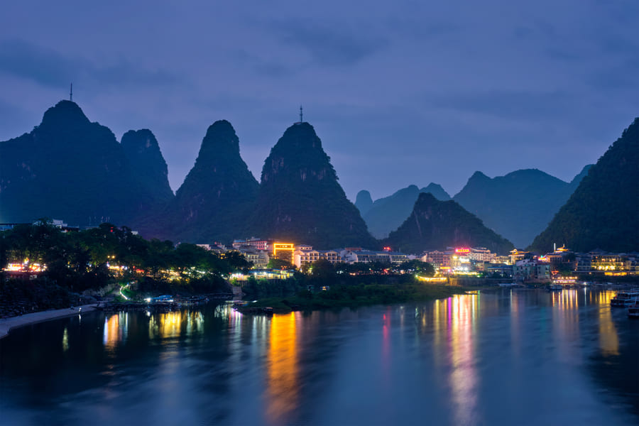 Vista nocturna de un río tranquilo que refleja las luces brillantes de un pueblo asentado al pie de imponentes montañas kársticas, bajo un cielo crepuscular azul oscuro.