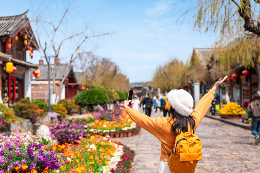 Una turista con mochila amarilla y gorro blanco se toma una foto con su celular en una colorida calle peatonal de un pueblo tradicional chino.